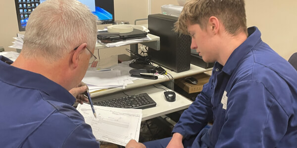 Two men in blue work uniforms look over paperwork at a desk with a computer and office stationery.