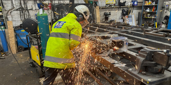 Apprentice in a hi-vis jacket using an angle grinder on metal, sending out sparks in an industrial workshop.